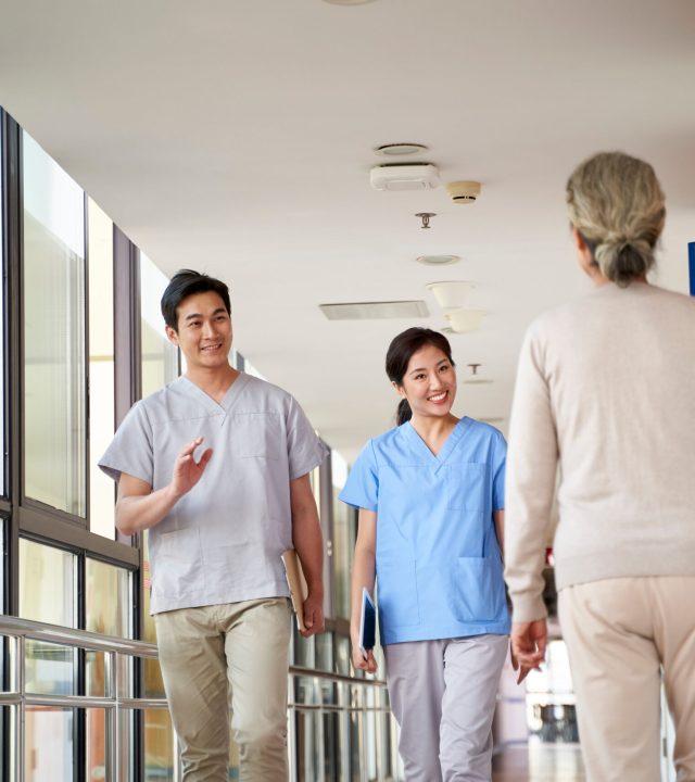 Happy,Young,Asian,Physical,Therapists,Greeting,Residents,In,Hallway,Of Happy,Young,Asian,Physical,Therapists,Greeting,Residents,In,Hallway,Of